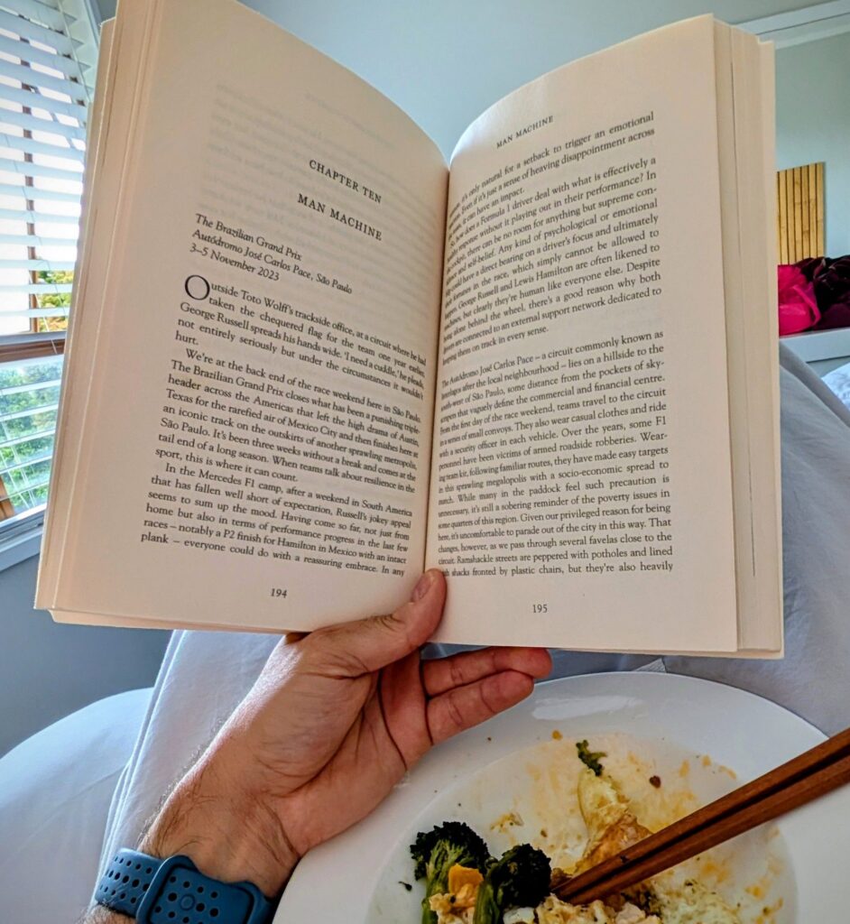 Perspective of reading a book in bed with a plate of food below and light coming from the window